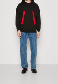 Felpa nera con strisce di accento rosse e tasca frontale, abbinata a jeans di denim blu. Presenta una vestibilità rilassata e un cappuccio con coulisse. Scarpe nere.