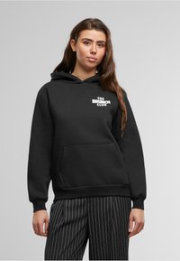 Black hoodie made of cotton blend, featuring a front pocket and white text "THE BRUNCH CLUB" on the chest. Smooth texture, relaxed fit.