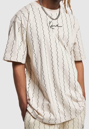 Personne portant un t-shirt blanc ample avec des lignes noires ondulées verticales et un petit logo noir sur la poitrine, associé à un pantalon assorti.