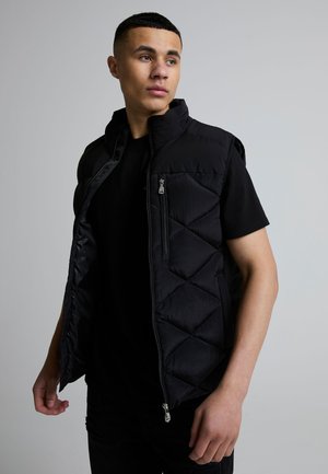 Ung mann iført svart polstret vest uten ermer over svart t-skjorte, ser til siden mot en ensfarget grå bakgrunn.