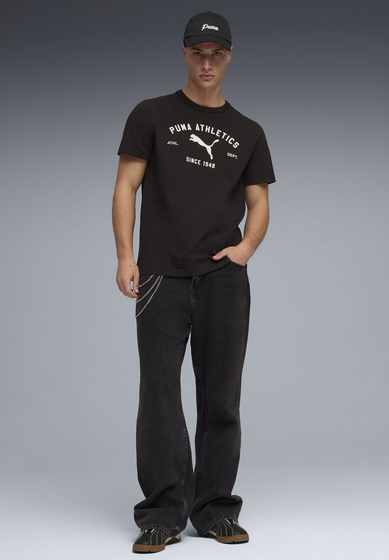 T-shirt en coton noir avec le logo "PUMA Athletics", jean en denim noir avec accent chaîne argentée, casquette de baseball noire avec logo, et chaussures vertes.