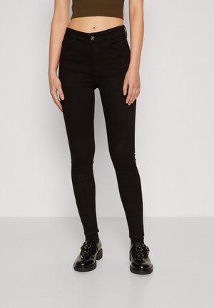 Kobieta w czarnych obcisłych dżinsach z wysokim stanem, brązowym krótkim topie i czarnych błyszczących sznurowanych butach, stojąca na jednolitym tle.