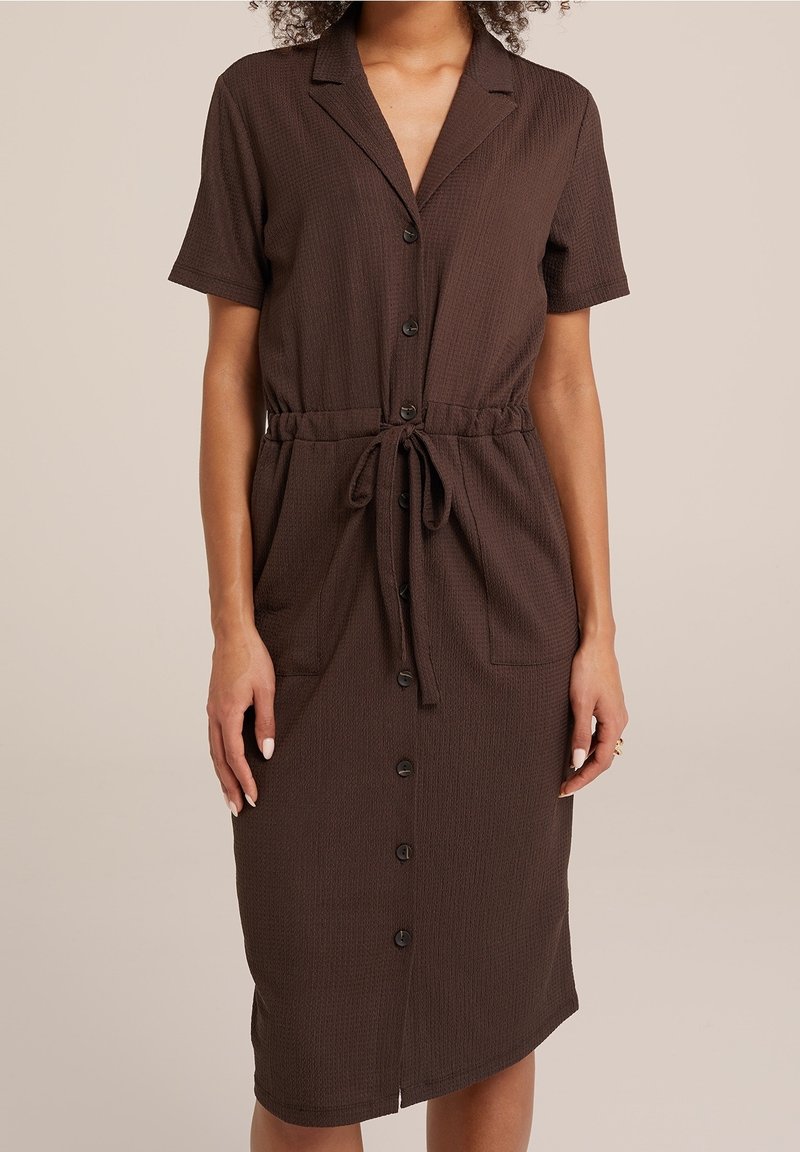 Brun kortärmad klänning i strukturerad tyg, knäppning fram och knytmidja. Har två sidofickor och en medellång längd.