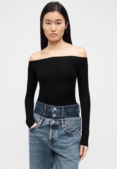 Μαύρο κορμάκι με μακριά μανίκια και άνοιγμα στον ώμο, σε συνδυασμό με μπλε ψηλόμεσο τζιν παντελόνι με πολλαπλά κουμπιά και ξεθωριασμένη υφή.