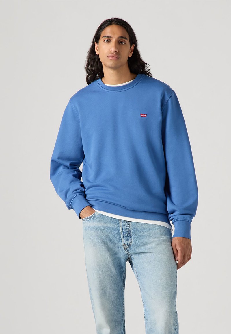 Blue sweatshirt made of soft fabric, featuring a round neckline and ribbed cuffs. Small red logo emblem on the chest. Worn with light denim jeans.