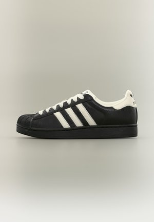 Fekete alacsony szárú sportcipő fehér Adidas csíkokkal, fehér fűzőkkel és texturált orrmellel egyszínű háttéren.