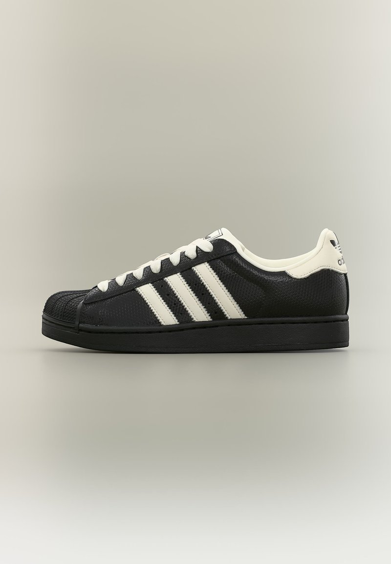 Μαύρο αθλητικό παπούτσι χαμηλού ύψους με λευκές ρίγες Adidas, λευκά κορδόνια και υφασμένη μύτη σε μονόχρωμο φόντο.