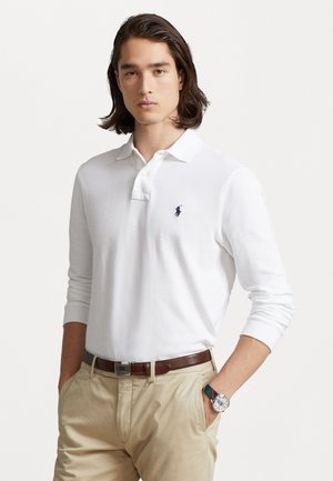 Weißes Langarm-Polohemd mit blauem gesticktem Logo, kombiniert mit beigen Hosen und einer mehrfarbigen Uhr. Glatte Stoffoberfläche.