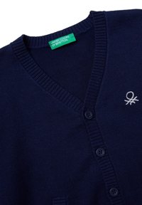 Námořnicky modrý pletený kardigan s žebrovaným výstřihem do V, zapínáním na knoflíky, malým bílým vyšívaným logem a zelenou štítkem „United Colors of Benetton“ uvnitř límce.