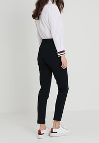 Svarta slim-fit byxor med hög midja, slät textur, i kombination med en vit skjorta med knappar och avslappnade vita sneakers med röda detaljer.
