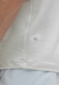 Svetlo šedé športové tričko s hladkým, textúrovaným materiálom; obsahuje malé logo na leme a zaoblený spodný okraj.