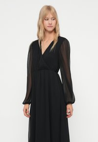 Frau mit blonden Haaren, die ein langes schwarzes Kleid mit durchsichtigen langen Ärmeln trägt, steht vor einem einfachen hellen Hintergrund und schaut zur Seite.