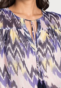 Blusa multicolore con scollatura a V e laccetto con ponpon, realizzata in tessuto leggero. Il design presenta un motivo a zigzag in viola, giallo e beige.