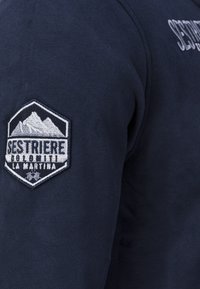 Mörkblå sweatshirt med en hexagonal broderad patch med bergsmotiv, med texten "Sestriere" och "La Martina." Tillverkad i bomull med texturerad yta.
