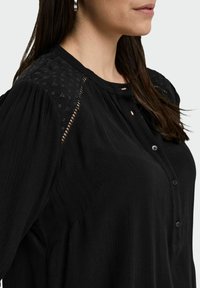 Blusa nera in tessuto texturizzato, con fronte bottonato, cuciture decorative sulle spalle e maniche leggermente a sbuffo.