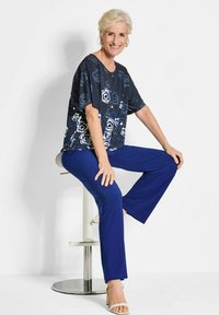 Marineblauw bloemenprinttop met een losse fit en korte mouwen, gecombineerd met effen blauwe rechte broek. Model zit op een barkruk.