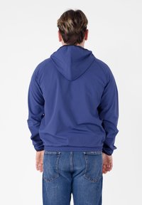 Giacca con cappuccio blu navy, con polsini e orlo elasticizzati, realizzata in materiale liscio. Cuciture visibili e design semplice, indossata con jeans in denim.