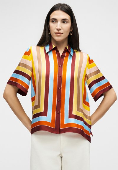 Γυναίκα με μακριά σκούρα μαλλιά, φοράει κοντομάνικο πολύχρωμο πουκάμισο με ρίγες και λευκό παντελόνι, στέκεται με τα χέρια στη μέση.