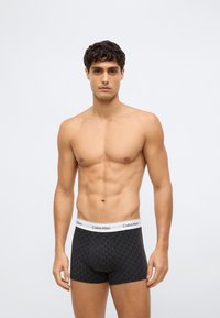 Muž bez trička s krátkymi kučerami, odetý v čiernych vzorovaných boxerkách Calvin Klein, stojí pred jednoduchým svetlým pozadím.