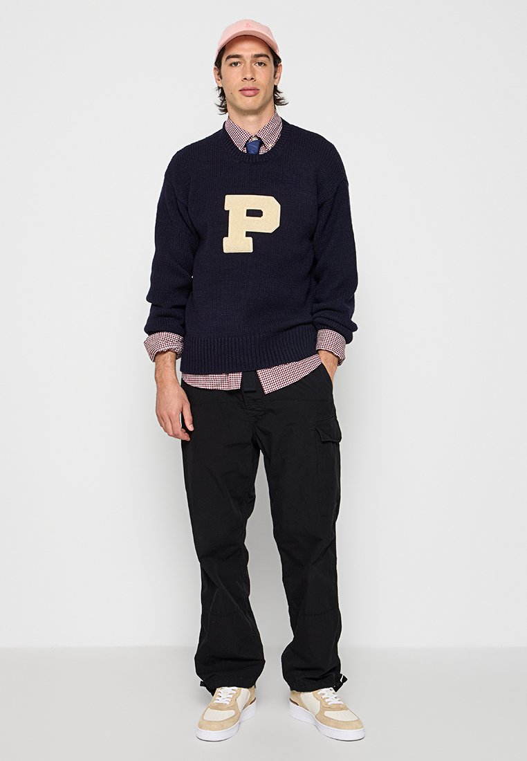 Navy gestrickter Pullover mit übergroßem beigefarbenem "P"-Logo, kombiniert mit einem karierten Hemd. Schwarze Cargo-Hosen und leichte Sneaker runden den Look ab.