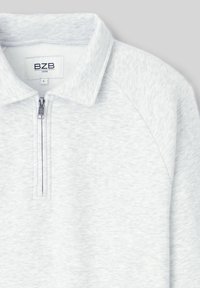 Pull-over gris clair à quart de zip avec col, fermeture éclair visible et étiquette de marque BZB à l'intérieur du col.