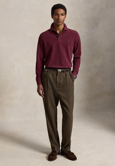 Homme portant un polo à manches longues bordeaux, un pantalon à carreaux marron taille haute, des chaussures habillées marron et une montre, debout avec une main dans la poche.
