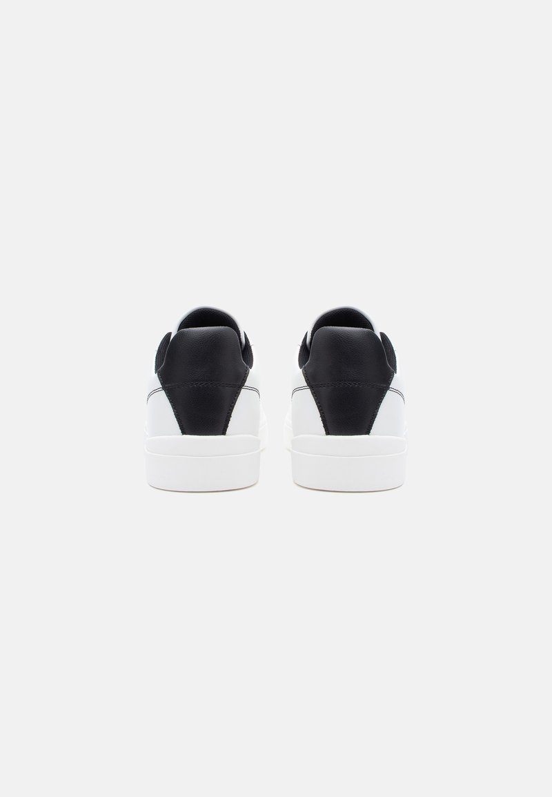Zapatillas de cuero en blanco y negro con un acabado liso, que cuentan con un talón acolchado y un diseño clásico. Estética minimalista, suelas planas.
