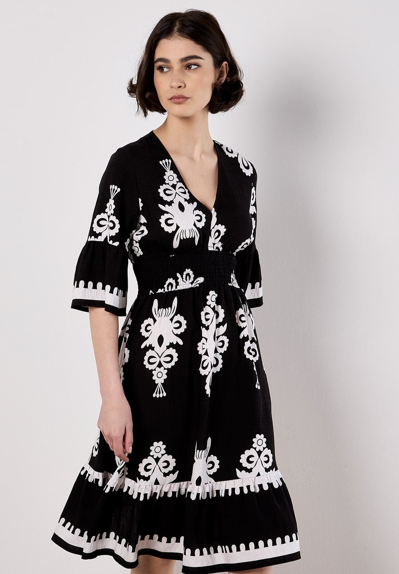 Le modèle porte une robe noire à motifs floraux blancs et une jupe évasée, se tenant devant un fond uni, regardant sur le côté.