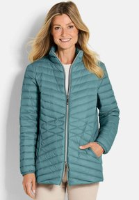 Smiling woman wearing a teal quilted jacket with a front zipper and side pockets, standing against a plain light background.
