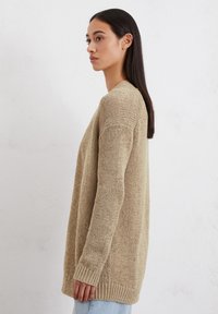 Beige gebreide cardigan met een losse pasvorm, lange mouwen en geribbelde zoom. De textuur is chunky, met een casual, open ontwerp.