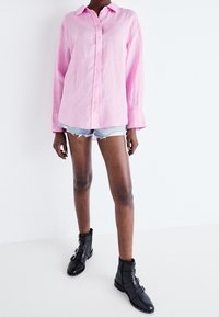 Persona che indossa una camicia a maniche lunghe rosa chiaro con bottoni, pantaloni corti in denim azzurro sfilacciati e stivaletti neri con fibbie.