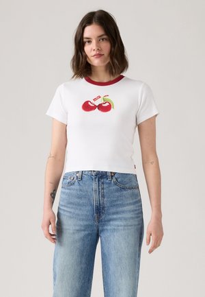 Femeie cu păr închis la culoare până la umeri poartă un tricou scurt alb cu guler roșu și grafică cu cireșe, asortat cu blugi albaștri cu talie înaltă.