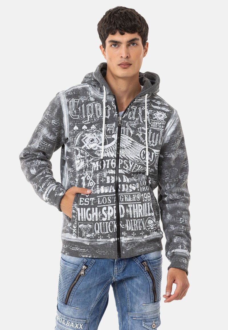 Grauer Reißverschluss-Hoodie mit grafischen Drucken, Kordelzugkapuze und Fronttaschen. Kombiniert mit blauen Jeanshosen mit Reißverschlusdetails.