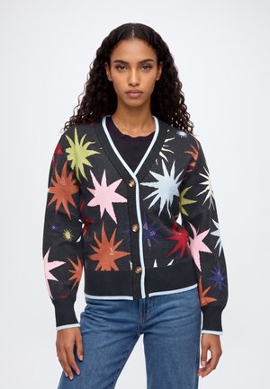 Un mannequin porte un cardigan noir à motifs colorés en forme d’étoiles, un haut noir, un jean bleu et des chaussures à talons, et tient un sac à main noir.