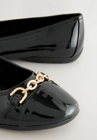Ballerines en vernis noir avec un bout rond et une accent en chaîne dorée à l'avant. Texture lisse avec un talon légèrement rehaussé.