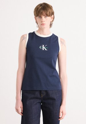 Marineblauw mouwloos topje met een ronde witte halslijn, voorzien van een groen en witte "CK"-logo in het midden. Draagt bij donkere blauwe jeans.