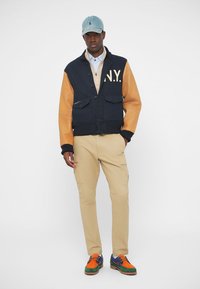 Στρατιωτικό μπλε και καφέ τζάκετ με γράμματα "N.Y.", μπεζ πουλόβερ από κάτω, χακί παντελόνι και πολύχρωπα παπούτσια. Χαλαρή, στιβαρή εμφάνιση.