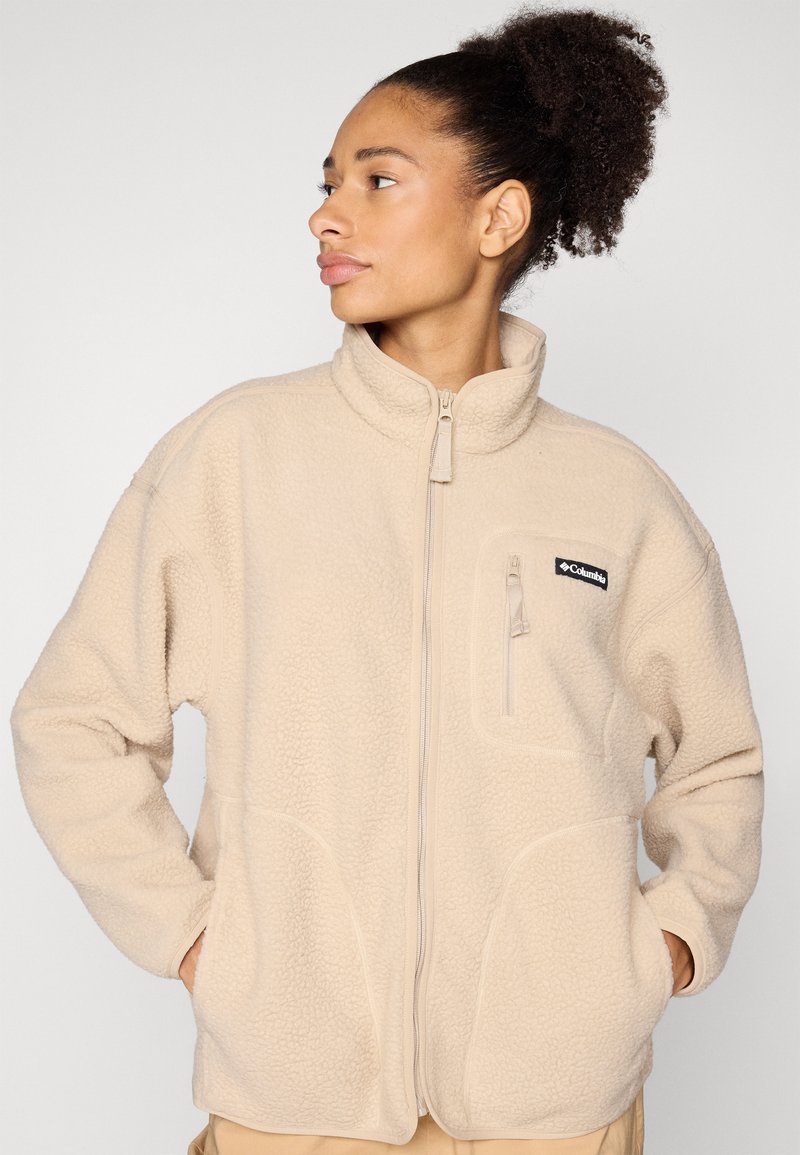 Frau mit lockigem, zurückgebundenem Haar, die eine beige Fleecejacke trägt und die Hände in den Taschen hat, schaut seitlich vor einem einfarbigen Hintergrund.