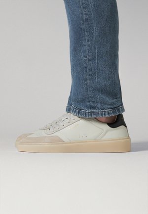 Persoon die een blauwe spijkerbroek en crèmekleurige sneakers draagt met beige suède accenten en een zwarte hielband, staand op een lichtgrijze ondergrond.