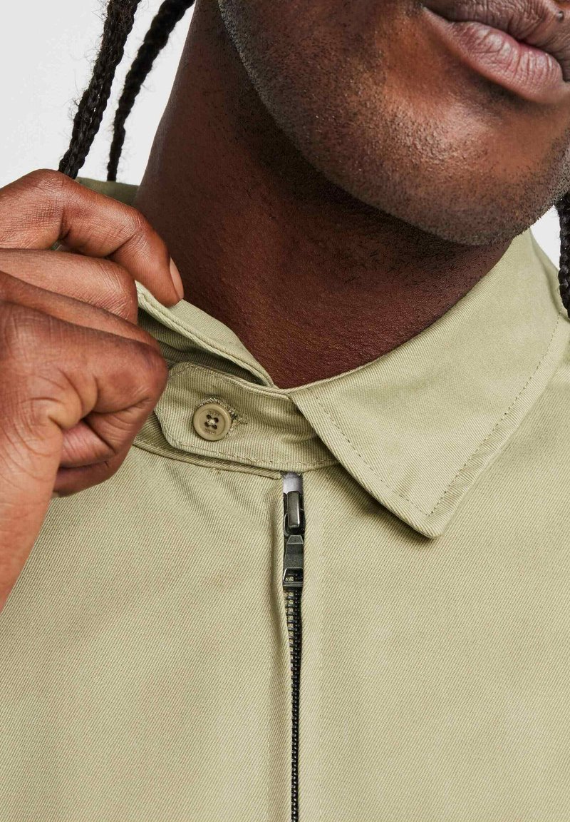 Chaqueta color caqui claro con cierre delantero, textura tejida y un cuello abotonado con un acabado suave. Primer plano de los detalles de la tela y el hardware.