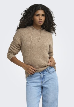 Femme aux cheveux bouclés portant un sweat à capuche en maille marron et un jean bleu clair, posant avec une main sur la hanche et l'autre tenant son pull.