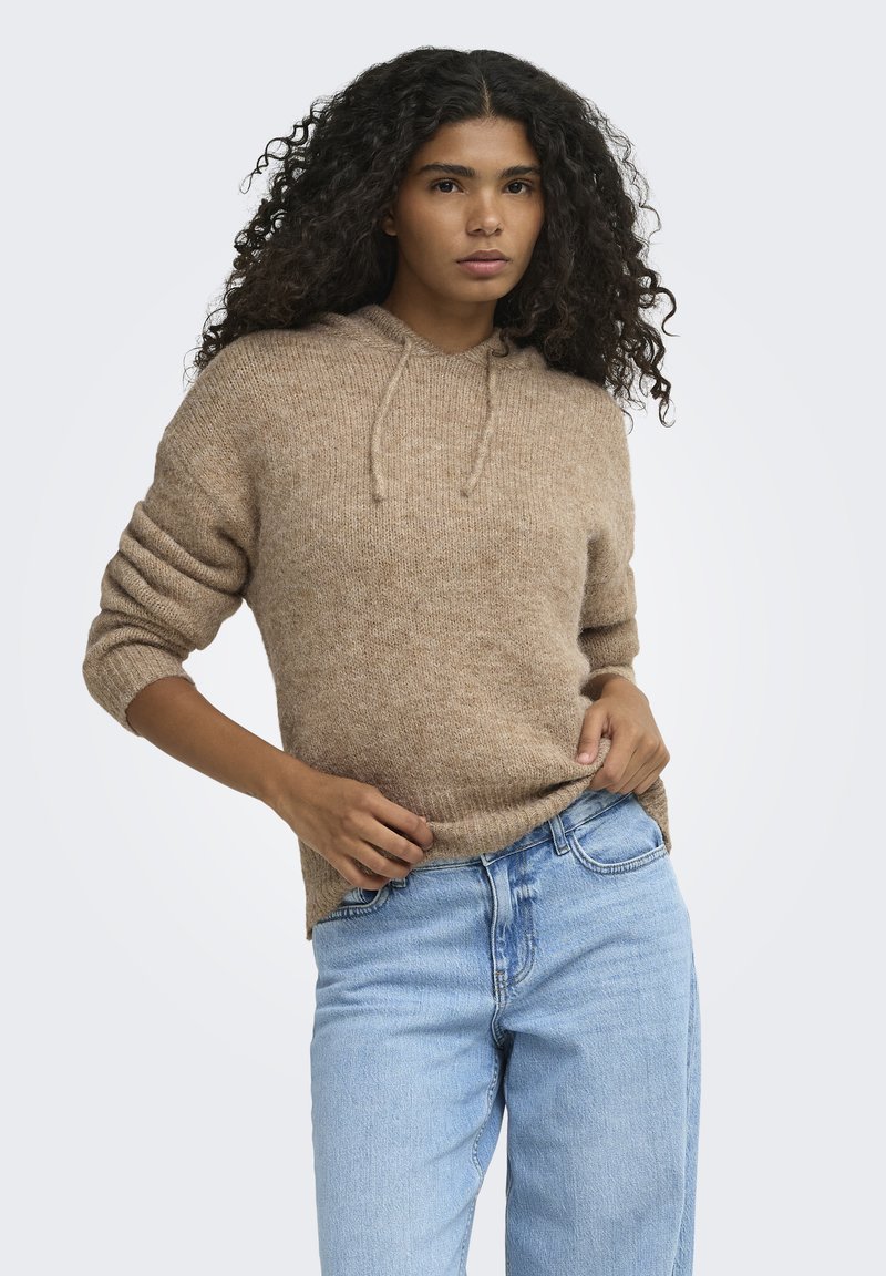 Femme aux cheveux bouclés portant un sweat à capuche en maille marron et un jean bleu clair, posant avec une main sur la hanche et l'autre tenant son pull.