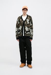 Pletena kardigan s kamuflažnim uzorkom u smešnoj, crnoj i beloj boji, s V izrezom, crnim dugmadi i dva prednja džepa. Uparen s tamnim cargo hlačama i bež čizmama.