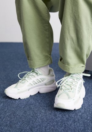 Personne portant un pantalon vert clair et des baskets blanches avec des touches vertes, debout sur un sol moquetté bleu.