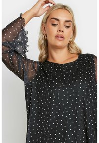 Top nero con pois bianchi, con maniche trasparenti e arricciate e una vestibilità comoda. Tessuto liscio, scollatura rotonda e stile casual.