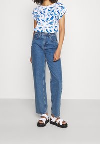 White short-sleeve shirt with blue bird print, paired with high-waisted blue jeans and white flat sandals with black soles.