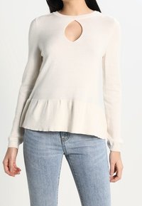 Sweter w kolorze kremowym z długim rękawem, posiadający dekolt w kształcie dziurki i peplum, wykonany z miękkiego materiału, charakteryzujący się gładką teksturą oraz dopasowanymi rękawami.