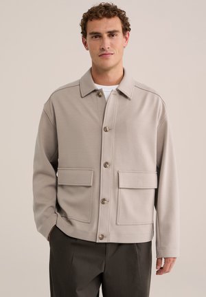 Homme portant une veste beige boutonnée avec de grandes poches avant et un pantalon foncé, debout devant un fond clair uni.
