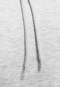 Šedá mikina z látky s blízkým záběrem na splétané šňůrky. Měkká textura s jemným melírováním a plastovými hroty na koncích.