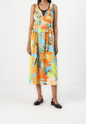 Femme portant une robe midi sans manches à imprimé tropical coloré avec une ceinture à nouer à la taille et des chaussures plates noires, debout devant un fond blanc uni.